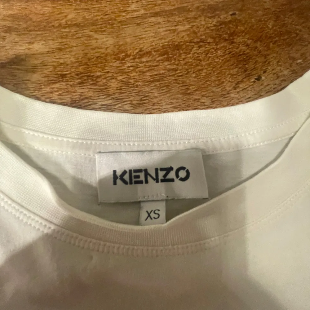 Säljer en snygg vit t-shirt från Kenzo i storlek Xs som är väldigt liten i storleken. Den har ett coolt tigertryck i mitten med texten 'Kenzo Paris' i guld och mörkblått. Perfekt för en casual look eller att styla upp med en kavaj. T-shirten är i bomull och har korta ärmar. Passar perfekt till både jeans och kjol!. T-shirts.