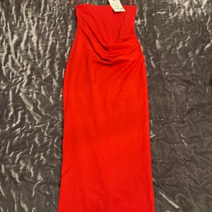 Röd klänning med smala axelband - Snygg röd klänning i ett mjukt material. Perfekt för festliga tillfällen! Klänningen har en elegant drapering framtill och är i ett nytt skick med prislappen kvar. Passar perfekt för en kväll ute eller en speciell middag.
