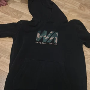 Svart hoodie från WRP - Säljer en svart hoodie från WRP med coolt camouflagemönster och texten 'Unperfect Antics' på bröstet. Perfekt för en avslappnad stil. Den har en stor ficka fram och en bekväm huva. Passar perfekt för höst och vår!