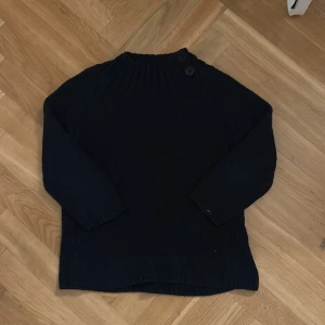 Mörkblå stickad tröja med trekvartsärmar - Säljer en mörkblå stickad tröja med ribbad krage och två knappar vid halsen. Perfekt för vintern. Tröjan har trekvartsärmar och är i bra skick. Passar både till vardags och lite finare tillfällen.