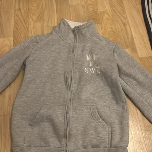 Grå fleecejacka från Rosetti - Mysig grå fleecejacka från Rosetti med dragkedja och hög krage. Perfekt för kyliga dagar och ridning. Jackan har broderad text 'SWE' på bröstet och är i mycket bra skick. Passar perfekt för höst och vår!