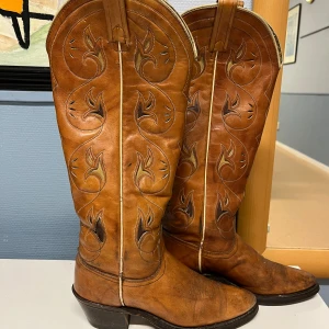 Bruna cowboy boots i skinn - Säljer ett par snygga bruna cowboy boots i skinn med coola broderade mönster. De har en robust klack och är i bra skick, perfekta för både vardag och fest. Sulorna är svarta och ser slitstarka ut. Passar perfekt för höst och vår!