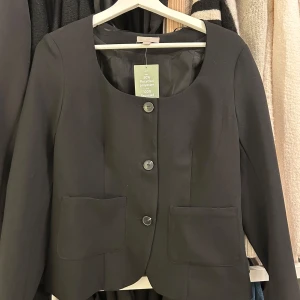 Svart kavaj med knappar - Snygg svart kavaj i klassisk stil med stora knappar framtill. Den har en rundad hals och två praktiska fickor. Perfekt för både vardag och festliga tillfällen. Materialet är 100% polyester vilket gör den lätt och bekväm att bära.