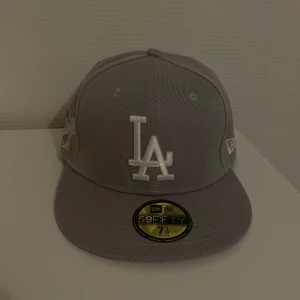 Grå keps från New Era, LA keps - Snygg grå keps från New Era med LA-logga framtill och World Series-märke på sidan. Kepsen är i 59FIFTY-modell och har en platt skärm. Perfekt för att ge din outfit en sportig touch!