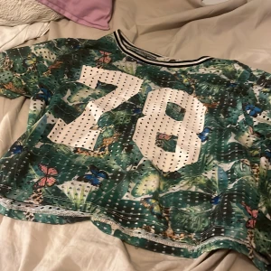 Grön mönstrad topp från H&M - Säljer en cool grön topp från H&M med ett tropiskt mönster av fjärilar och växter. Den har en stor vit siffra '78' på framsidan och är kortärmad med en croppad passform. 