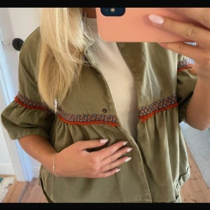 Trekvartsärmad jacka  - Säljer en superfin olivgrön jacka med broderade detaljer i rött och svart. Jackan har trekvartsärmar och en loose passform, perfekt för vår och höst. Den är i mycket bra skick och har en unik stil med knappar framtill. Passar perfekt till både vardag och fest!