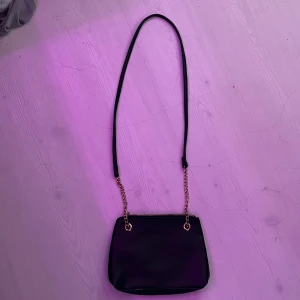 Svart axelväska med guldkedja - Snygg svart axelväska i skinn med guldkedjedetaljer. Perfekt för både vardag och fest. Väskan har en dragkedja upptill för säker förvaring av dina saker. Den är stilren och lätt att matcha med olika outfits.