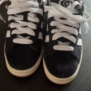 Svarta Adidas Campus sneakers - Säljer ett par svarta Adidas Campus sneakers i bra skick. De har en klassisk design med vita ränder och snörning. Skorna är i mocka och har en bekväm passform. Perfekta för vardagsbruk och ger en stilren look.