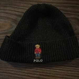 Svart ribbad mössa från Polo Ralph Lauren - Säljer en snygg svart ribbad mössa från Polo Ralph Lauren. Den har en broderad nalle i röd tröja och texten 'POLO' framtill. Perfekt för att hålla sig varm och stilren under kalla dagar. Passar de flesta då den är i one size. Vill också till lägga att den är lite nopprig 
