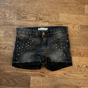 Svarta jeansshorts med nitar - Snygga svarta jeansshorts med coola nitar framtill. De har en fransig kant nertill och klassisk femficksdesign. Perfekta för en avslappnad sommarlook eller en festival. Passar till allt från t-shirts till blusar!
