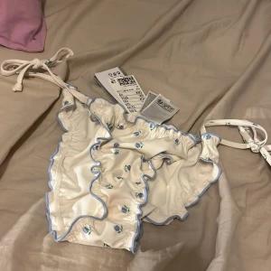 Vit blommig bikiniunderdel från H&M - Säljer en superfin vit bikiniunderdel från H&M med blå blommor och volangdetaljer. Den har knytband på sidorna för justerbar passform. Perfekt för sommarens stranddagar! 🌸