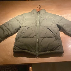 Olivgrön pufferjacka från Dickies - Säljer en olivgrön pufferjacka från Dickies i mycket bra skick. Jackan har en dragkedja framtill och är perfekt för kalla dagar. Den är vadderad och har långa ärmar med elastiska muddar för extra värme. Insidan är fodrad med Dickies-logga. Perfekt för höst och vinter! Använde den förra vintern men är nu för liten. Men jag använde inte den så mycke så den är i väldig bra skick. Pris kan diskuteras!
