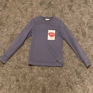 Mörkblå tröja från Limitato - Säljer en snygg mörkblå tröja från Limitato med ett coolt tryck av röda läppar på bröstet. Tröjan har långa ärmar och är perfekt för en avslappnad stil. Passar både till vardags och fest. Skicka ett meddelande vid intresse!