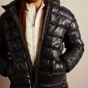 Svart Glansig pufferjacka - Säljer en snygg svart pufferjacka i glansigt material. Jackan har en dragkedja framtill och långa ärmar med ribbade muddar. Perfekt för kalla dagar och håller dig varm och stilren. Passar både till vardags och lite finare tillfällen.