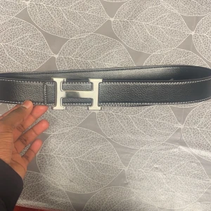 Svart bälte med H-spänne - Säljer ett stilrent svart bälte med ett stort H-spänne i silver. Bältet är i skinn och har en klassisk design som passar perfekt till både vardag och fest. Det är i mycket bra skick och har en elegant look. Perfekt för att ge din outfit det där lilla extra! 🖤