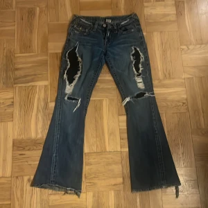 Blå bootcut jeans med slitningar - Säljer ett par blå bootcut jeans från True Religion i bra skick. De har coola slitningar på framsidan och en snygg detalj med slitna kanter vid bensluten. Perfekta för en avslappnad och trendig look. Passar bra till både vardag och fest!