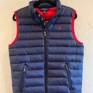 Blå dunväst från Polo Ralph Lauren - Säljer en snygg blå dunväst från Polo Ralph Lauren med röd insida och detaljer. Västen har dragkedja och två fickor med dragkedjor. Perfekt för höst och vår, både stilren och praktisk. Passar lika bra till vardags som till lite finare tillfällen.