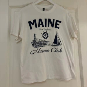 Vit t-shirt med marintryck - Säljer en vit t-shirt med tryck från 'Maine Marine Club'. Den har ett coolt marint tema med en fyr och segelbåt. T-shirten är kortärmad och i bomull, använt 2 gånger, därav mycket bra skick!