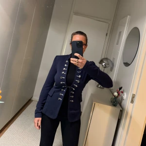 Mörkblå kavaj med dekorativa knappar - Snygg mörkblå kavaj i stretch från Fifth Avenue med dekorativa silverknappar framtill. Perfekt för både vardag och fest. Den har en figurnära passform och långa ärmar. Kavajen ger en stilren och elegant look.