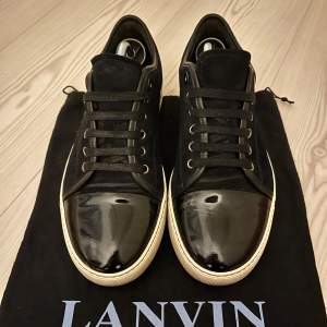 Snygga svarta sneakers från Lanvin i skinn med glansig finish på tån. De har en klassisk snörning och en vit sula som ger en stilren kontrast. Perfekta för både vardag och festliga tillfällen. Skorna är i mycket bra skick och kommer med Dustbag