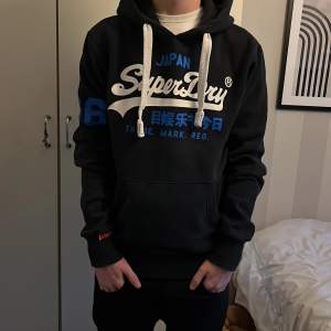 Säljer en svart hoodie från Superdry i mycket bra skick. Den har en stor logga i vitt och blått på framsidan och siffran 66 i blått på ärmen. Hoodien har en huva med tjocka vita snören, Perfekt för höst och vinter. passa på och köp!