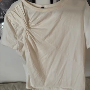Beige t-shirt från Divided - Säljer en snygg vit t-shirt från Divided i storlek L. Den har en unik rynkdetalj på ena axeln som ger en cool twist.