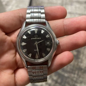 Eterna-Matic Kontiki herrklocka - Säljer en klassisk Eterna-Matic Kontiki herrklocka med en stilren design. Klockan har en svart urtavla med datumvisning och silverfärgade timmarkeringar. Boetten och armbandet är i silverfärgat stål, vilket ger en tidlös och elegant look. Perfekt för både vardag och fest!