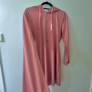 Rosa långklänning med huva - Säljer en superfin rosa långklänning med huva. Den har långa ärmar och en avslappnad passform. Klänningen är gjord i ett mjukt material som känns skönt mot huden.