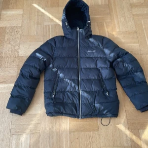 Svart dunjacka från GANT - Säljer en snygg och varm svart dunjacka från GANT. Jackan har en praktisk dragkedja och två fickor med dragkedjor. Perfekt för kalla vinterdagar med sin vadderade design och huva som skyddar mot vind och kyla. Den är i nyskick och passar 170-180cm
