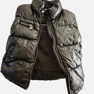 Svart dunväst från H&M med Hello Kitty - Säljer en superfin svart pufferväst från H&M med ett gulligt Hello Kitty-märke på bröstet och texten 'Hello Kitty' på ryggen. Perfekt för att hålla sig varm och stilren under kalla höstdagar. Fint skick. Tvättas innan det skickas. Storlek: 122