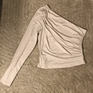Beige oneshoulder topp från H&M - Säljer en stilren beige oneshoulder topp från H&M. Toppen har en lång ärm och en elastisk midja för en bekväm passform. Perfekt för en trendig look.