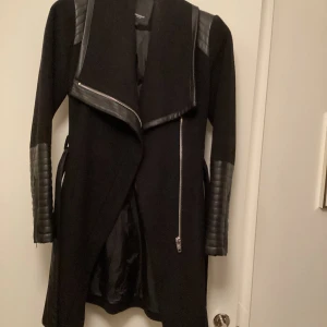 Svart kappa från Rockandblue - Snygg svart kappa från Rockandblue med coola detaljer i skinn på axlar och ärmar. Jackan har en asymmetrisk dragkedja och en stilren design som passar perfekt för höst och vinter. Perfekt för dig som vill ha en edgy look!