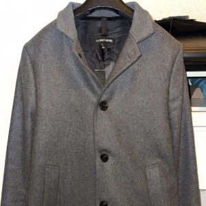 Helt ny grå kappa från J.Lindeberg Holger 2.0 - Hej. Säljer denna fina J.Lindeberg Holger 2.0 kappan som är helt ny oanvänd. Storlek 50 (M/L). Alla etiketter samt prislappen sitter kvar. Kappan är köpt på J.Lindeberg, kvittot finns. Säljes då den är för liten för mig.