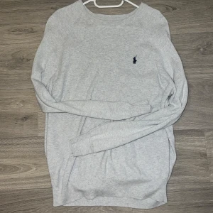Ralph Lauren tröja - Säljer en stilren grå tröja från Ralph Lauren med rund hals och broderad logga på bröstet i ny skick. Perfekt för både vardag, skjorta under och fest. Storlek M men passar L med. 