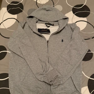 Grå hoodie från Ralph Lauren - Säljer en stilren grå hoodie från Ralph Lauren med dragkedja och huva. Tröjan har en liten broderad logga på bröstet och är tillverkad i mjuk bomull. Perfekt för en avslappnad look. Medium men passar lika bra för small.
