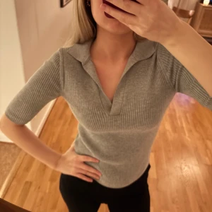 Grå ribbad polotopp - Säljer en stilren grå ribbad polotopp med korta ärmar. Perfekt för en avslappnad men ändå chic look. Toppen har en klassisk krage och en smickrande passform.