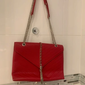 Röd axelväska med kedjedetaljer - Snygg röd axelväska i syntetmaterial med quiltad design och silverfärgade kedjor. Väskan har en elegant framsida med en dekorativ kedja och tofsdetalj. Perfekt för att ge en färgklick till din outfit.