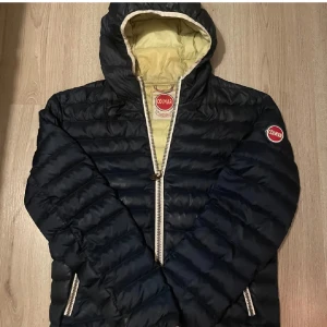  pufferjacka från Colmar - Snygg svart pufferjacka från Colmar med beige foder och huva. Jackan har en dragkedja framtill och Colmar-logga på bröstet och ärmen. Perfekt för kyliga dagar. Lite missfärgning som man kan se på bilden men är bara det på insidan