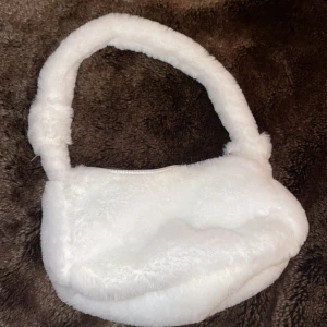 Vit fluffig axelväska - Säljer en supermjuk och fluffig vit axelväska med dragkedja. Perfekt för att ge din outfit en mysig touch. Väskan har ett litet och endå rätt rymligt fack och en bekväm axelrem. Aldrig använd. Guldig dragkedje grej men inte själva dragkedjan. 