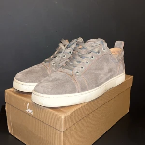 Grå sneakers från Christian Louboutin - Snygga grå sneakers i mocka från Christian Louboutin med den ikoniska röda sulan. Skorna har snörning och en stilren design som passar perfekt för en trendig look.