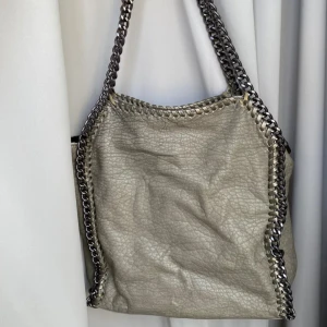 Beige axelväska med kedjedetaljer - Snygg beige axelväska med grova kedjedetaljer längs kanterna och axelremmen. Väskan har en dragkedja för säker förvaring och är tillverkad i ett skinnliknande material med en texturerad yta.