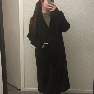 Svart kappa från Noa Noa - Elegant svart kappa från Noa Noa med klassisk design. Kappan har långa ärmar och knäpps med knappar framtill. Perfekt för kyligare dagar med sin stilrena look.