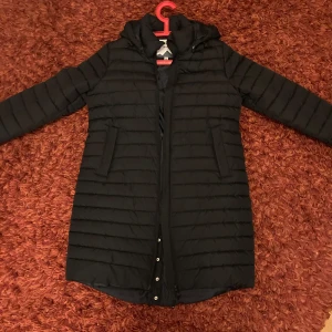 Svart lång kappa med huva  - Snygg svart lång kappa med huva och knappar där bak och är perfekt för kyligare dagar. Den quiltade designen ger extra värme och stil.