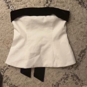 Vit tubtopp med svart rosett - Elegant vit tubtopp med en stor svart rosett baktill. Toppen har en stilren design och är perfekt för festliga tillfällen.