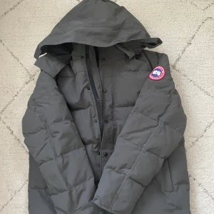 Svart dunjacka från Canada Goose - Säljer en svart dunjacka från Canada Goose med huva och det klassiska märket på ärmen. Jackan är perfekt för kalla vinterdagar och har en stilren design med dragkedja och knappar framtill. Helt bekväm och skön att ta på sig inför vintern just nu. Storlek är XL men passar också M och L.