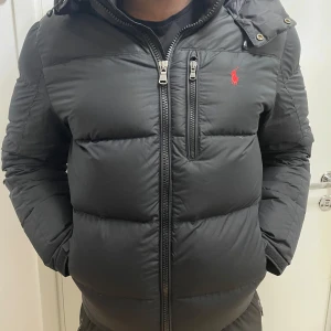 Svart dunjacka från Ralph Lauren - Snygg svart dunjacka från Ralph Lauren med luva och dragkedja. Jackan har en broderad logga på bröstet och praktiska fickor. Perfekt för kyliga dagar. Jackan har ett få tal skador vid ärmen