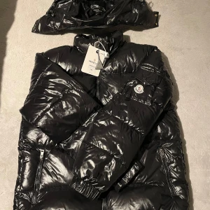 Svart pufferjacka från Moncler - Snygg svart pufferjacka från Moncler med avtagbar huva och dragkedja. Jackan har ett stilrent märke på ärmen och är perfekt för kalla dagar.