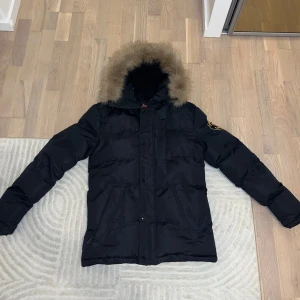 Svart dunjacka från Zavetti Canada - Snygg svart dunjacka från Zavetti Canada med pälskantad huva. Jackan har långa ärmar och stängs med dragkedja och knappar. Perfekt för kalla vinterdagar. Är inte säker om det är en kill eller tjej modell men den funkar till båda, sitter lite mer oversized på tjejer. Pris kan diskuteras