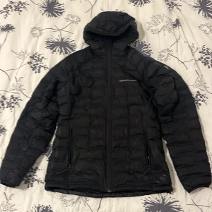 Svart dunjacka från Peak Performance - Säljer en svart dunjacka från Peak Performance. Jackan har en quiltad design med dragkedja och huva. Perfekt för kyliga dagar. Priset kan diskuteras.