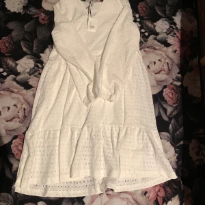 Vit klänning med volangdetaljer - Charmig vit klänning med volangdetaljer och broderat mönster. Klänningen har en luftig passform och är perfekt för varma dagar. Den är ärmlös och har en vacker struktur som ger en romantisk känsla.